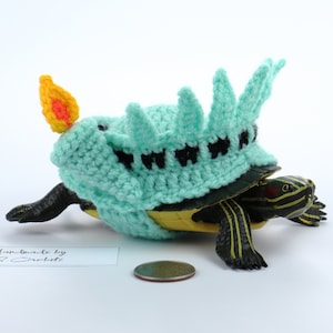 May include: A small turtle wearing a crocheted Statue of Liberty costume. The costume is green and white with a black crown and a yellow flame on top. The turtle is sitting on a white surface with a US quarter coin in front of it. Handmade by S. Crochet.