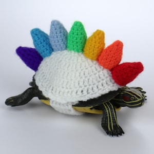 May include: A small turtle wearing a crocheted rainbow-colored shell with a white crocheted top.