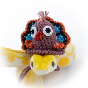 May include: A crochet turkey hat featuring a brown base, a red wattle, an orange beak, and a colorful fan-shaped tail with blue, black, and orange yarn accents. The hat is topped with a small tuft of white, pink, and blue yarn fibers.