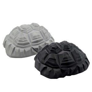 May include: Two geometric turtle shells, one in light gray and one in black. Each shell features a faceted, multi-layered design with distinct angular plates and a textured surface, presented as separate decorative objects without any visible base.