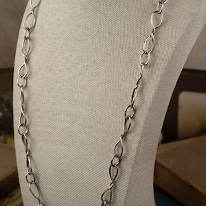 May include: A silver-toned necklace with a long chain of interconnected oval and teardrop-shaped links. The necklace is displayed on a neutral-colored bust, showcasing its design and length. The jewelry piece is a simple yet elegant accessory.
