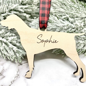 May include: A wooden Christmas ornament shaped like a dog with the name "Sophie" engraved on it. The ornament is attached to a red and black buffalo plaid ribbon.
