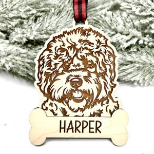 Puede incluir: Un adorno navideño de madera con la cara de un perro y el nombre "HARPER" grabado en una etiqueta con forma de hueso.