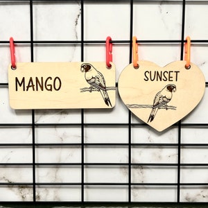 May include: Two wooden birdcage name tags, one rectangular with the word "MANGO" and a parrot, the other heart-shaped with the word "SUNSET" and a parrot.