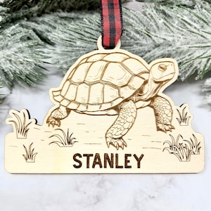 May include: A wooden Christmas ornament featuring a laser-engraved illustration of a tortoise walking on grass. The ornament is personalized with the name "STANLEY".