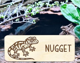 May include: A rectangular wooden sign with rounded corners. The sign features a detailed brown laser-cut gecko illustration on the left and the name "NUGGET" in brown capital letters on the right. The background includes greenery and wood.