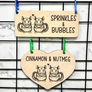 Pode incluir: Duas placas de madeira com ilustrações de chinchilas. A placa superior, retangular, diz "SPRINKLES & BUBBLES". A placa inferior em forma de coração diz "CINNAMON & NUTMEG". Ambas as placas mostram um par de chinchilas e estão penduradas numa grelha preta.