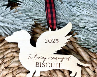 Könnte beinhalten: Holz-Dackel-Ornament mit Engelsflügeln, dem Jahr 2025 und dem Text "In loving memory of BISCUIT". Der Schmuck hängt an einem rot-schwarz karierten Band, vor dem Hintergrund von gefrorenem Grün und gewebtem Material.