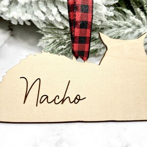 May include: A wooden cat ornament with the name "Nacho" engraved on it. The ornament is painted in a light wood color and has a red and black buffalo plaid ribbon attached.