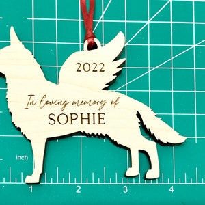 German Shepherd with angel wings memorial ornament. Ornament is made of wood with, “In loving memory of”, dog’s name, and year engraved on it. Ornament is hung with red ribbon and about 4.25 inches wide and 3.22 inches tall.