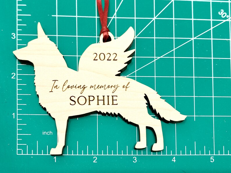 German Shepherd with angel wings memorial ornament. Ornament is made of wood with, “In loving memory of”, dog’s name, and year engraved on it. Ornament is hung with red ribbon and about 4.25 inches wide and 3.22 inches tall.
