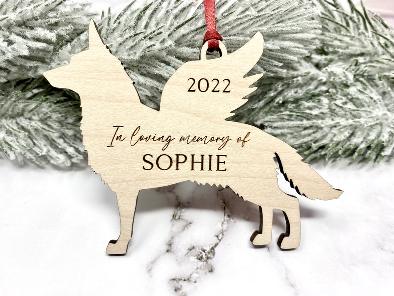 German Shepherd with angel wings memorial ornament. Ornament is made of wood with, “In loving memory of”, dog’s name, and year engraved on it. Ornament is hung with a red ribbon.
