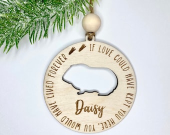 Könnte beinhalten: Weihnachtsbaumschmuck aus Holz mit einem ausgeschnittenen Meerschweinchen und dem Text "Wenn Liebe dich hier hätte behalten können, hättest du für immer gelebt." Der Schmuck enthält auch zwei kleine Pfotenabdrücke und den Namen "Daisy".