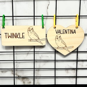 May include: Two wooden birdcage name tags, one rectangular with the name "TWINKLE" and the other heart-shaped with the name "VALENTINA". Both tags feature a laser-engraved image of a parakeet.