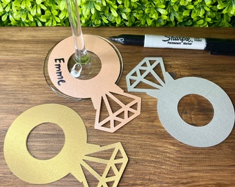 May include: Three gold, silver, and rose gold ring-shaped wine glass charms with diamond cutouts. The rose gold charm has the name "Emme" written on it.