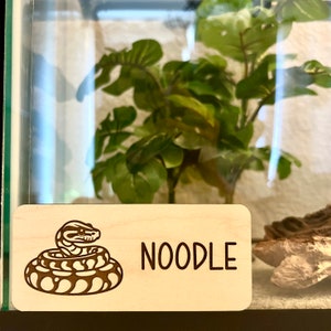 May include: A wooden sign with a black and white illustration of a snake curled up. The sign says "NOODLE" in black text.