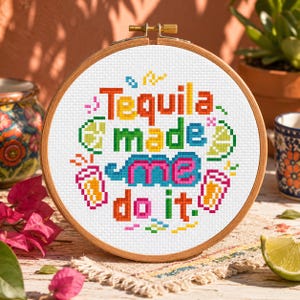 Könnte beinhalten: Ein runder Holzstickrahmen mit weißem Stoff und einem Kreuzstichmotiv. Der Text "Tequila made me do it" ist in bunten Blockbuchstaben gestickt, umgeben von gestickten Symbolen von Limettenspalten und zwei Gläsern mit einer goldfarbenen Flüssigkeit.
