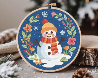 May include: A cross-stitch embroidery hoop featuring a snowman design. The snowman wears an orange hat and scarf, surrounded by a floral wreath and snowflakes on a blue background. The wooden hoop is visible.