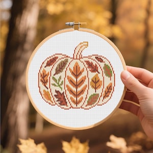 May include: A cross-stitch pumpkin design in a wooden embroidery hoop. The pumpkin is filled with autumn leaves in shades of orange, brown, and green. The background is a blurred view of a forest with fall foliage.