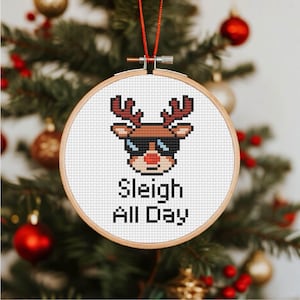 Könnte beinhalten: Ein runder, kreuzgestickter Weihnachtsschmuck mit einem pixeligen Rentier mit Sonnenbrille. Der Text "Sleigh All Day" steht unter dem Rentier. Der Schmuck hängt vor einem verschwommenen Weihnachtsbaumhintergrund mit roten und goldenen Ornamenten.