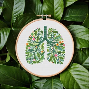 May include: A cross-stitch embroidery of lungs, filled with green leaves and small orange and pink flowers, set in a wooden hoop. The design is on a white background, surrounded by large green leaves.