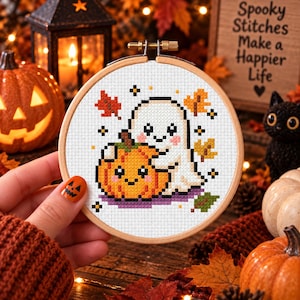 May include: A circular wooden embroidery hoop containing a cross-stitch pattern of a white ghost hugging an orange pumpkin. The design includes small orange and red autumn leaves, purple ground details, and tiny yellow stars on a white fabric background.