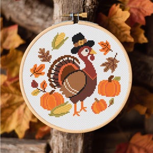 May include: A cross-stitch embroidery in a wooden hoop features a Thanksgiving turkey wearing a pilgrim hat, surrounded by pumpkins, corn, and fall leaves. The color palette includes shades of brown, orange, and green, creating a festive autumn theme.