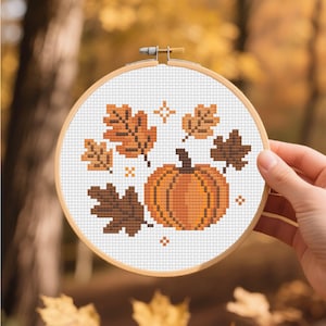 May include: A cross-stitch embroidery hoop with a pumpkin and autumn leaves design. The pumpkin is orange, and the leaves are shades of brown. The embroidery is on a white background, framed by a wooden hoop.