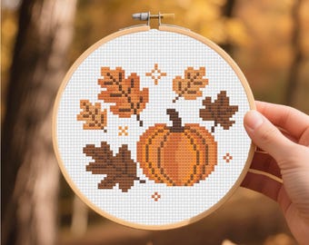 May include: A cross-stitch embroidery hoop featuring a pumpkin and fall leaves design. The pumpkin is orange, and the leaves are various shades of brown. The embroidery is set against a white background, framed by a wooden hoop.