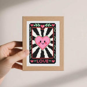 Könnte beinhalten: Gerahmtes Kreuzstich-Kunstwerk mit einem rosa Herz mit lächelndem Gesicht, vor einem schwarz-weißen Sonnenstrahl-Design. Das Wort "LOVE" ist unten gestickt. Der Rahmen besteht aus hellfarbenem Holz.