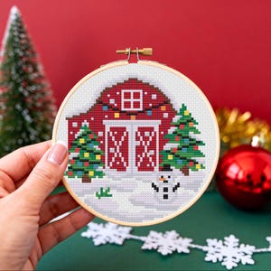 May include: A cross-stitch hoop featuring a red barn with white doors, a small window, and Christmas lights. Two green Christmas trees flank the barn, and a snowman stands in the snow. The hoop is held by a hand.