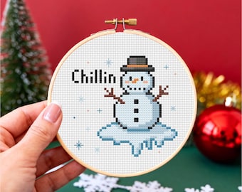 May include: A cross-stitch embroidery hoop featuring a pixelated snowman design. The snowman wears a black hat and has the word "Chillin" above it. The design is set against a white background with blue snowflakes.