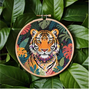 Può includere: Ricamo a punto croce di una faccia di tigre, in un cerchio di legno. La tigre ha strisce arancioni e nere, con motivi floreali e di foglie colorati. Lo sfondo è verde scuro.