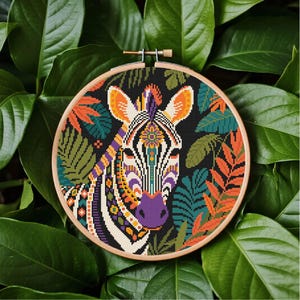 Op de afbeelding: Een kleurrijk kruissteekborduurwerk van een zebra gezicht in een houten hoepel. De zebra heeft een patroon met oranje, paarse en gele accenten. De achtergrond is zwart met groene en oranje bladeren.