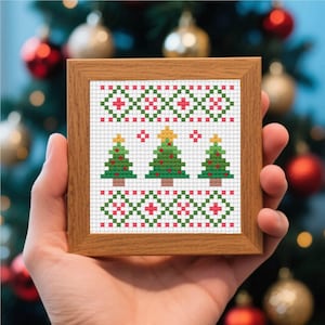 May include: A framed cross-stitch featuring three green Christmas trees with yellow star toppers, set against a white background. The design includes red and green geometric patterns. The wooden frame is square.