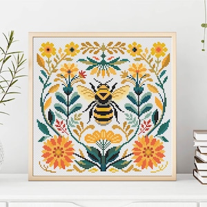 May include: A framed cross-stitch featuring a large bee in the center, surrounded by yellow and orange flowers, green leaves, and a light beige background. The artwork is in a wooden frame and sits on a white surface.