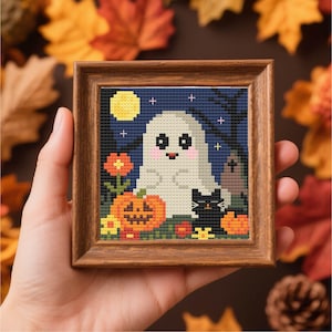 May include: A framed cross-stitch featuring a Halloween scene. The artwork depicts a friendly ghost, a black cat, and a carved pumpkin under a full moon. The background is a dark blue night sky with a few cross-stitched trees and a tombstone.