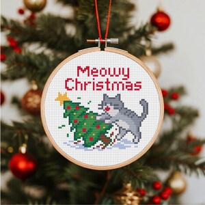 Può includere: Ornamento natalizio rotondo a punto croce, in un cerchio di legno. Il design presenta un gatto grigio e un albero di Natale verde con decorazioni rosse. Il testo "Meowy Christmas" è rosso.