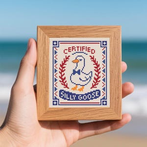 Pode incluir: Uma pequena moldura de madeira contendo um bordado em ponto cruz de um ganso branco com uma gravata borboleta azul. O texto diz CERTIFIED SILLY GOOSE, rodeado por ramos vermelhos e uma borda geométrica azul e vermelha em tecido branco.