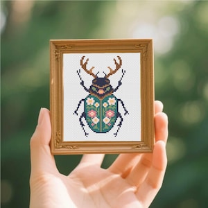 May include: A small, rectangular, light brown wooden frame containing a cross-stitch embroidery of a stag beetle. The beetle has brown antlers, a dark head, and a teal body decorated with small pink and white flowers on a white fabric background.