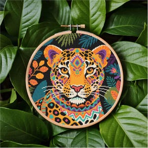 May include: A colorful cross-stitch embroidery of a jaguar's face, set in a wooden hoop. The jaguar features orange, yellow, purple, and teal patterns. The background is black with green and orange foliage. The hoop is surrounded by large green leaves.