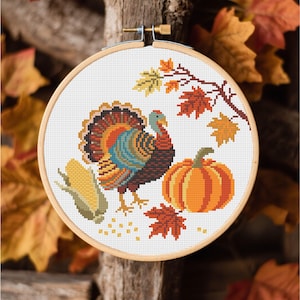 May include: A cross-stitch embroidery hoop featuring a colorful turkey, pumpkin, and corn. The design includes fall leaves in shades of orange, yellow, and red. The wooden hoop is set against a rustic wooden background, evoking a harvest theme.
