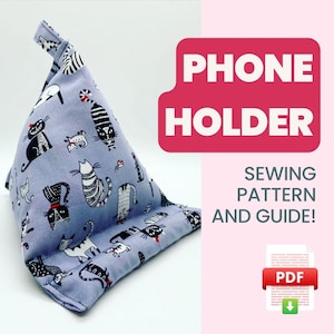 以下が含まれることがあります： 猫柄の紫色の携帯電話ホルダー。ホルダーは三角形で、持ち手があります。画像の右側に「PHONE HOLDER SEWING PATTERN AND GUIDE! PDF」というテキストがあります。