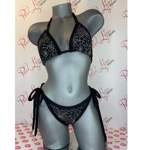 以下が含まれることがあります： 白いクモの巣のデザインが施された黒いビキニ。トップは三角形スタイルで、ボトムはチークカットです。