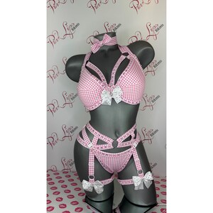 May include: A pink and white gingham lingerie set displayed on a mannequin. The set comprises a halter top, choker, high-waisted bottom, and garter straps. White bows with polka dots adorn the lingerie. A repeating logo forms the background.