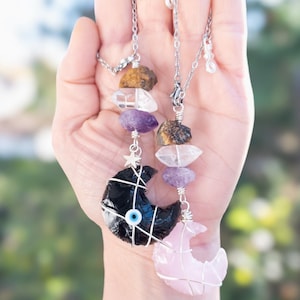 May include: Two silver chain necklaces with crystal and gemstone pendants. One necklace features a black crescent moon with an evil eye charm and the other features a pink crescent moon. Both necklaces have a star charm.
