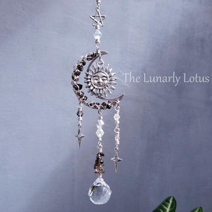 May include: A celestial-themed suncatcher featuring a silver crescent moon with a smiling sun face, adorned with crystals and stars. A clear crystal ball hangs below. The Lunarly Lotus is printed on the image.