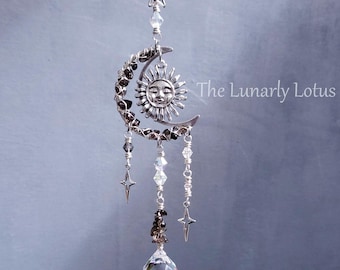 May include: A celestial-themed suncatcher featuring a silver crescent moon with a smiling sun face, adorned with crystals and stars. A clear crystal ball hangs below. The Lunarly Lotus is printed on the image.