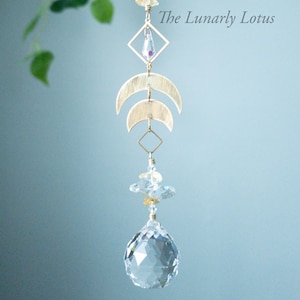 May include: A gold crystal suncatcher with a crystal ball at the bottom. The suncatcher features a gold chain, a cluster of clear and gold crystals, a gold diamond shape, and two gold crescent moons. The text "The Lunarly Lotus" is visible in the image.