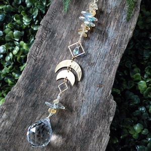 May include: A crystal suncatcher with gold accents and a clear crystal ball hanging from a wooden board. The suncatcher features a geometric design with crescent moons and a small crystal in a square frame. The board is surrounded by green foliage.